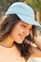 Close-up of tender young caucasian woman looks away smiles and collects her hair outdoor. Brunette female wears top and cap. Concept of vacation with great mood.