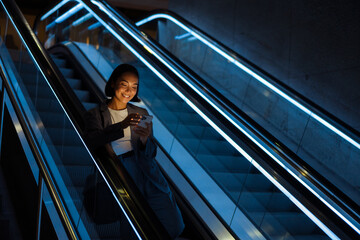 Young asian woman using mobile phone while standing on escalator indoors