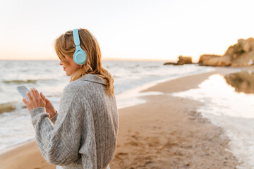 Beautiful woman listening music with headphones and mobile phone while standing at sunny beach