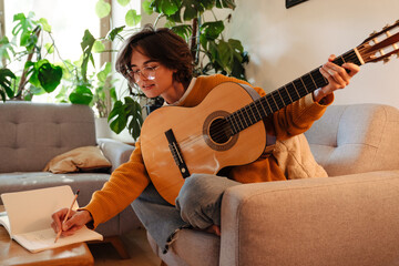 Brunette young woman playing guitar while sitting on armchair