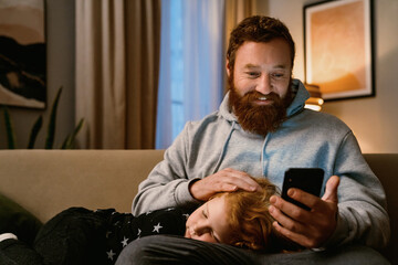 Smiling father using smartphone while resting with his son on couch