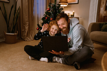 Father and son having video call via laptop while sitting in living room with Christmas tree on background