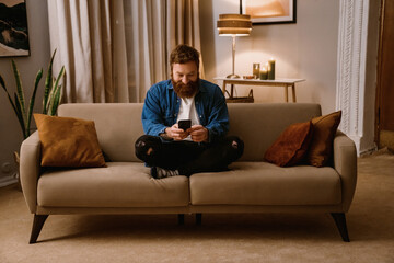Smiling bearded man using mobile phone while sitting in lotus pose on sofa at home