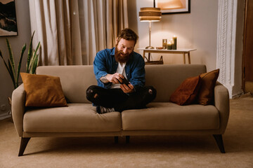 Fototapeta premium Excited man playing game on mobile phone while sitting on sofa at home