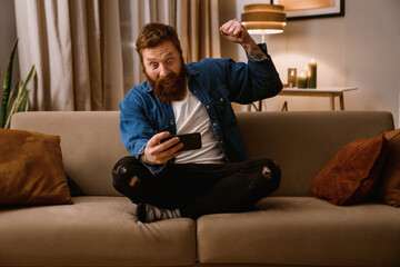 Obraz premium Excited bearded man playing game on mobile phone and showing winning gesture while sitting on sofa