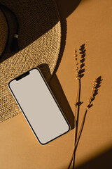 Luxury bohemian fashion online store, shop concept. Mobile phone, dried grass, straw hat on orange background with aesthetic sunlight shadows. Blank screen with mockup copyspace