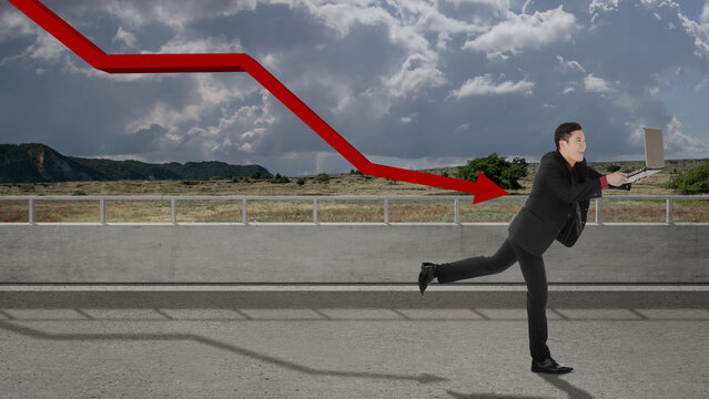 Asian businessman with a laptop running away from the decreased arrow