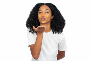 Fototapeta na wymiar Happy multiracial woman blowing kiss. Portrait of pleased young female model with dark curly hair in white T-shirt looking at camera with hand near mouth. Affection, flirting concept