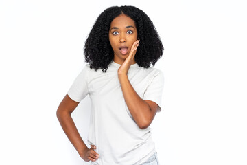 Fototapeta na wymiar Surprised African American woman pressing hand to cheek. Portrait of shocked young female model with dark curly hair in white T-shirt looking at camera with open mouth. Shock, gossip concept
