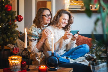 Smiling embracing happy lesbian couple in warm sweaters sitting on sofa at home near christmas tree and using smartphone - obrazy, fototapety, plakaty