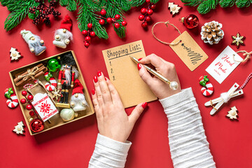 female hand holds pen and writing Christmas shopping list, gift ideas on white notepad on red background Top view Flat lay Holiday card Woman making Christmas presents list, Planning, buying concept