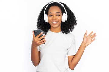 Portrait of happy young woman in headphones holding mobile phone. African American lady wearing white T-shirt listening to music and waving hand. Mobile technology and leisure concept