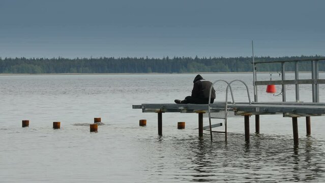 A man sitting on the ledge of the port in Estonia with the black coat and hat on him