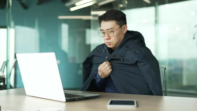 Cold asian businessman IT programmer suffers and trembles warms hands covering jacket try to work on laptop computer at desk in the modern office with problem of heating system indoors Low temperature