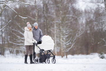Happy young family pushing baby stroller and walking on snow covered sidewalk at park in cold...