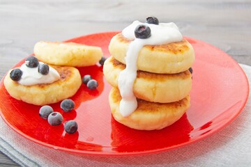 A stack of cottage cheese pancakes (syrnik) with sour cream and berries on a red plate. Delicious and healthy breakfast. Selective focus.