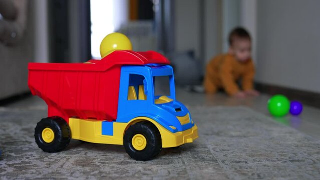 Colorful toy lorry on the floor of the room. Mom’s hand puts the plastic balls on the car. Crawling child appears at backdrop in blur.