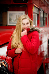 portrait of blonde beautiful smiling woman with long hair in red warm jacket staying near red coffee bus in winter park with snow