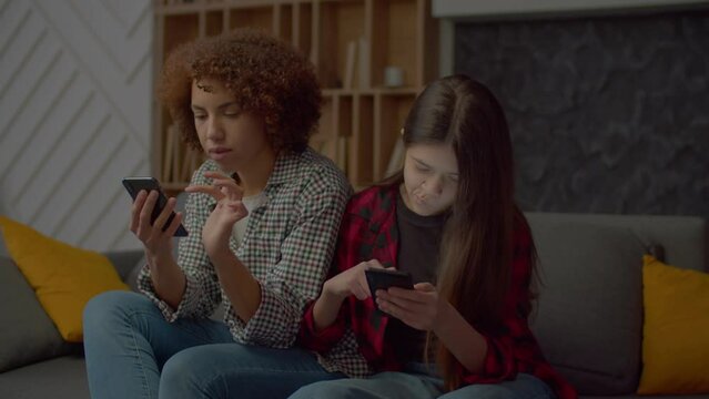Addicted to smartphones adorable multiracial teenage daughter and charming black mother phubbing each other, networking and browsing content online while sitting on sofa in domestic room.