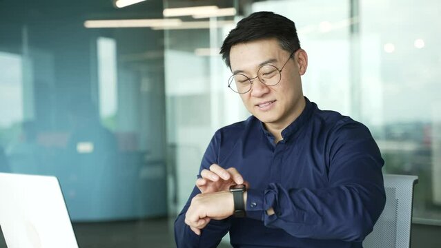 Smiling asian businessman scrolling on display smartwatch at modern glass workplace IT programmer entrepreneur using watch device gadget checking pulse using application touch screen online indoors