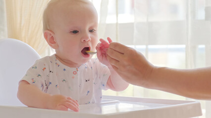 Mother is Feeding a baby from a spoon. Mom feeds a 9 month baby with fruit puree from a spoon, close-up, high key. Infant boy eats sitting on baby's chair. Mother cares about little son.