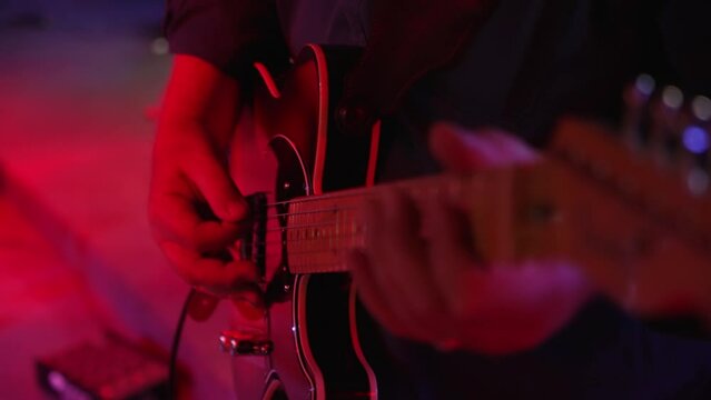 Male guitarist strums chords, plays electric guitar on stage lit in red lighting, slow motion close up 4K