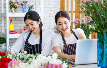 Millennial Asian young female flower shopkeeper decorator florist employee worker in apron sitting smiling writing checklist note while colleague typing laptop notebook computer in floral store