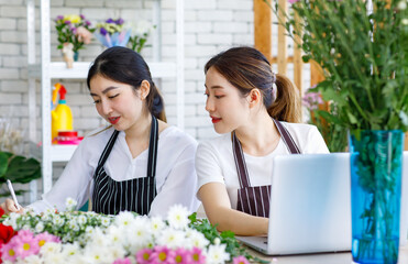 Millennial Asian young female flower shopkeeper decorator florist employee worker in apron sitting smiling writing checklist note while colleague typing laptop notebook computer in floral store