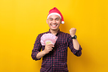 Excited young Asian man in Santa hat holding fan of cash money and raising fist, celebrating success on yellow studio background. Happy New Year 2023 celebration merry holiday concept