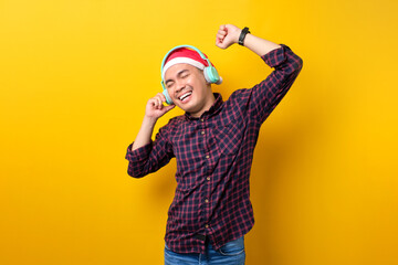 Fototapeta na wymiar Cheerful young Asian man in Santa hat listening music with headphones and dancing on yellow studio background. Happy New Year 2023 celebration merry holiday concept