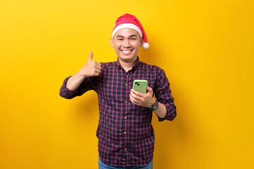 Fototapeta na wymiar Smiling young Asian man in Santa hat holding mobile phone and showing thumbs up over yellow studio background. Happy New Year 2023 celebration merry holiday concept