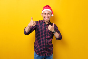 Cheerful young Asian man in Santa hat showing thumbs up, looking at camera on yellow studio background. Happy New Year 2023 celebration merry holiday concept