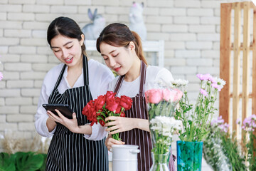 Millennial Asian young professional flower shopkeeper florist employee worker wearing apron standing smiling holding red roses bunch bouquet while colleague using tablet computer in floral store