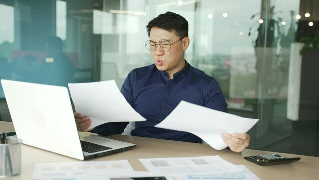Worried nervous asian businessman IT programmer having problems with paper work document at modern office Stressed man feeling exhausted with statistics reports analysing data at development company