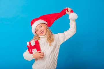 Fototapeta na wymiar A cute little girl in a Santa hat holds a gift on a blue background in the studio. Space for text. Winter concept, knitted sweater