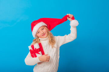 A cute little girl in a Santa hat holds a gift on a blue background in the studio. Space for text. Winter concept, knitted sweater