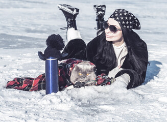 Fototapeta na wymiar Funny British fluffy cat and a girl in sunglasses and a bandana on their heads are fishing with winter fishing rods on the ice of the lake. Winter fishing with a cat. There is a thermos nearby.