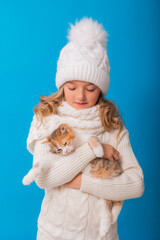 Obraz premium a cute little girl in a white knitted hat and sweater is holding a kitten on a blue background in the studio. Space for text. The concept of Christmas, the symbol of the year