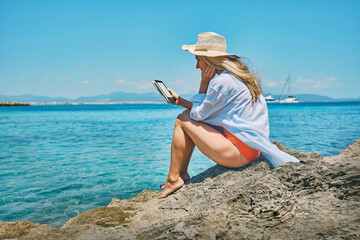 Fashion pretty woman outdoors lifestyle watching, reading on tablet ebook on the beach in summer day. Wearing wide brimmed hat, Sun bating with uv protection. Concept of beach vacation.
