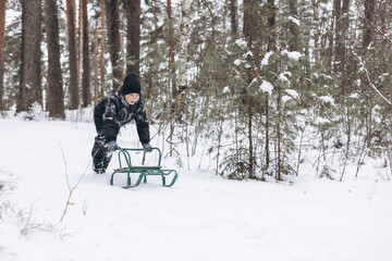 Happy teenager boy sledding and having fun outdoors. Joyful child playing in snow in winter forest. Laughing smiling kid walking in winter park in cold weather