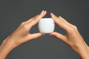 Human skincare beauty product. Studio photo of female hands is holding a white jar with cream against grey background. Daily care and hygiene routine.
