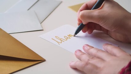 Close up of hands of woman writing a thank you note on a card, in golden calligraphy, expressing gratitude for a gift or service, with fancy golden and silver envelopes on her desk