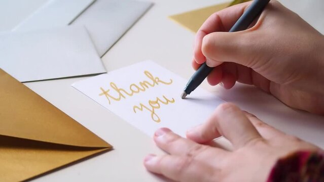 Close up of person writing thank you card by hand with calligraphy pen, in golden cursive letters. On the desk, fancy silver and gold envelopes are ready for sending a message of gratitude for a gift