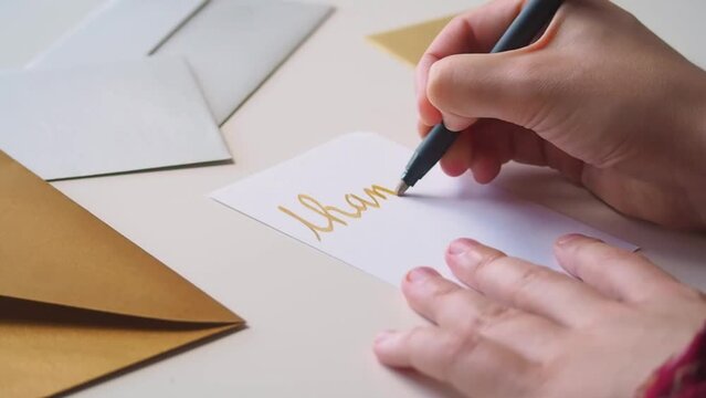Close up of hands of woman writing a thank you note on a card, in golden calligraphy, expressing gratitude for a gift or service, with fancy golden and silver envelopes on her desk