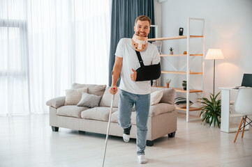 Fototapeta na wymiar Standing and smiling. Man with crutches is at home indoors. Having injuries