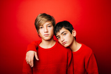 Studio portrait of two pre-adolescent boys, wearing a red sweater, embracing over a red background