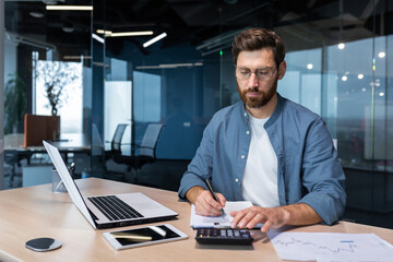 Serious and focused financier accountant on paper work inside office, mature man using calculator and laptop for calculating reports and summarizing accounts, businessman at work in casual clothes.