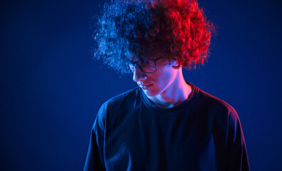 Looking down. Young man with curly hair is indoors illuminated by neon lighting