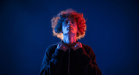 Confident look. Standing, serious facial expression. Young man with curly hair is indoors illuminated by neon lighting