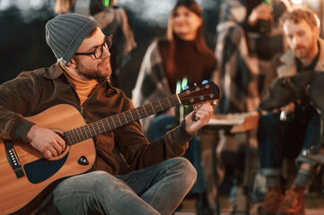 Obraz premium Man in hat is sitting and playing guitar. Group of people is spending time together on the backyard at evening time